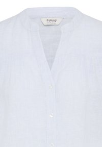 Blouse rayée bleu clair et blanc avec un col rond et de petits boutons blancs sur le devant, montrant l'étiquette de la marque b.young.