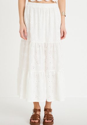 Kobieta ubrana w białą, haftowaną, warstwową spódnicę midi, zestawioną z brązowymi sandałami na paski i złotą bransoletą mankietową, na jednolitym tle.