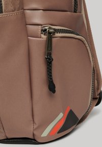 Zaino in tessuto beige con tasca frontale con zip, superficie testurizzata e dettagli decorativi in rosso, nero e grigio nell'angolo inferiore.