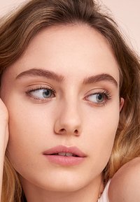 Närbild av ett ansikte med ljus hy. Drag inkluderar ljusbrunt hår, blågröna ögon, definierade ögonbryn och naturliga läppar. Bakgrunden är i mjuk fokus.