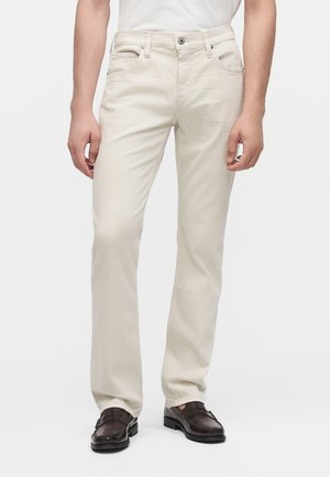 Man draagt off-white rechte broek en donkerbruine leren loafers, staat met ontspannen handen langs zijn zij.