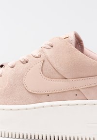 Světle růžová semišová teniska s bílou gumovou podrážkou, s prošitým logem Nike, zaoblenou špičkou a strukturovanými tkaničkami.