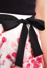 Cravate noire en tissu texturé, cintrant une jupe florale avec des motifs aquarelle roses et rouges sur un fond clair.