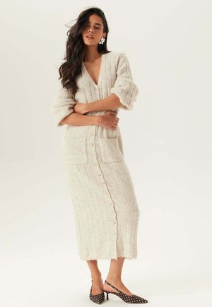 Kvinna med långt mörkt hår bär en krämfärgad stickad knappuppklänning med fickor och slingbacks med polkaprickar, står med armarna i kors.