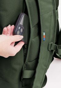 Zaino verde con tasca in rete, in fase di inserimento di uno smartphone. Presenta cerniere, spallacci regolabili e un piccolo accento multicolore.