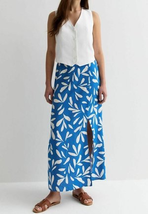 Femme portant un gilet blanc sans manches boutonné, une jupe maxi bleue avec des motifs de feuilles blanches et une fente à l'avant, ainsi que des sandales plates marron.