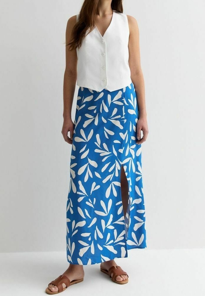 Femme portant un gilet blanc sans manches boutonné, une jupe maxi bleue avec des motifs de feuilles blanches et une fente à l'avant, ainsi que des sandales plates marron.