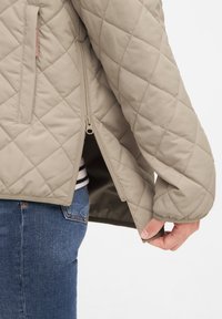 Beige gewatteerde jas met een ritszak aan de zijkant, gestructureerde stof en ribgebreide manchetten, gedragen over blauwe jeans.