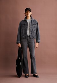 Zwarte denim jas met twee voorkruiken, grijs t-shirt, zwarte flared jeans, zwarte ballerina's en een zwarte baseballcap, terwijl een zwarte tote bag vasthoudt.