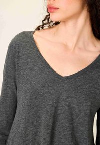 Moteris su garbanotais plaukais dėvi tamsiai pilką ilgomis rankovėmis su V formos iškirpte viršutinį rūbą, rodantį kaklą ir raktikaulius ant šviesaus fono.