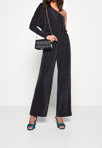 One-shoulder black jumpsuit with sparkly texture, wide legs, and a tied waist. Paired with a quilted black handbag and teal heeled sandals.