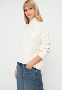 Pulover alb tricotat cu guler înalt, cu un model de tricotat în cablu și mansete cu țesătură în valuri, asociat cu blugi din denim albastri.