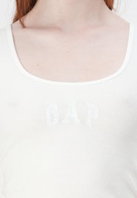 Balts ribots bez piedurknēm ar apaļu kakla izgriezumu un maigi iepresētu "GAP" logotipu uz krūtīm, demonstrējot gludu tekstūru.
