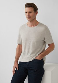 Helles graues, kurzärmliges Rundhals-T-Shirt aus weichem Stoff. Das Model trägt dunkle Jeans mit Händen in den Taschen und steht vor einem neutralen Hintergrund.