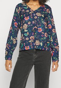 Blouse florale marine à col en V, ornée de fleurs et de feuilles colorées. Manches longues avec accents de boutons. Tissu lisse et léger.
