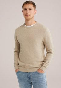 Beige kötött pulóver, textúrált felülettel, kerek nyakkivágással és bordázott mandzsettákkal. Világoskék farmernadrággal párosítva, kezek a zsebekben, semleges háttér.