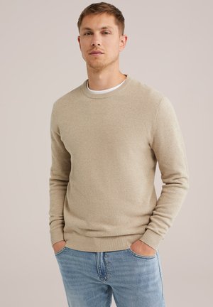 Suéter de punto beige con una superficie texturizada, escote redondo y puños acanalados. Combinado con jeans de color azul claro, con las manos en los bolsillos, fondo neutro.
