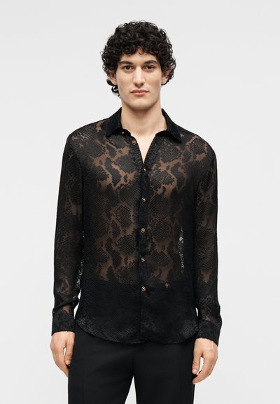 Homme aux cheveux bouclés portant une chemise noire transparente à boutons avec un imprimé animalier subtil et un pantalon noir, debout devant un fond uni.