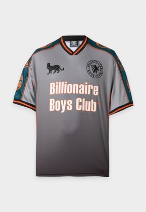 Mladý muž stojí s rukami za chrbtom, oblečený v sivé a čiernej tričku Billionaire Boys Club, svetlé rifle a čierno-biele tenisky.