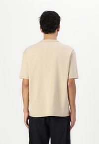 Beige kortærmet t-shirt med rund hals, der har en glat bomuldsstruktur og en afslappet pasform. Bagview viser lige søm og skuldersømme.