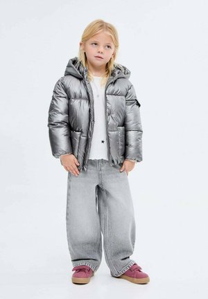 Niña pequeña con cabello rubio, vestida con una chaqueta acolchada plata, una camiseta blanca con estampado de estrellas, pantalones anchos grises y zapatillas rosas, de pie.