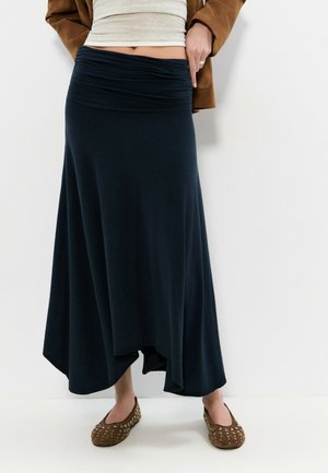 Femme portant une jupe asymétrique bleu marine, des ballerines beiges cloutées, un haut beige et une veste marron, debout contre un mur blanc.