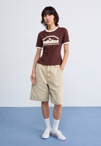Camiseta de algodão castanha com bordas brancas e logotipo da Adidas, combinada com calções de ganga bege. Modelo usa meias brancas e ténis com detalhes dourados.