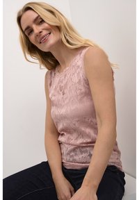 Rozā mežģīņu topiņš bez piedurknēm ar ziedu rakstiem, viļņotu kakla izgriezumu un pieguļošu siluetu, apvienots ar tumšiem džinsiem.