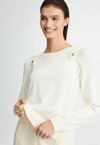 Pull en tricot couleur crème avec des détails côtelés aux manches et des accents de boutons dorés sur les épaules. Texture douce et coupe décontractée.