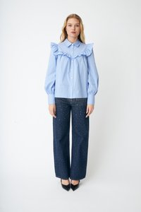 Kvinne iført en blåstripet bluse med volanger og vide, mørke jeans dekorert med små hvite prikker, stående foran en ensfarget hvit bakgrunn.