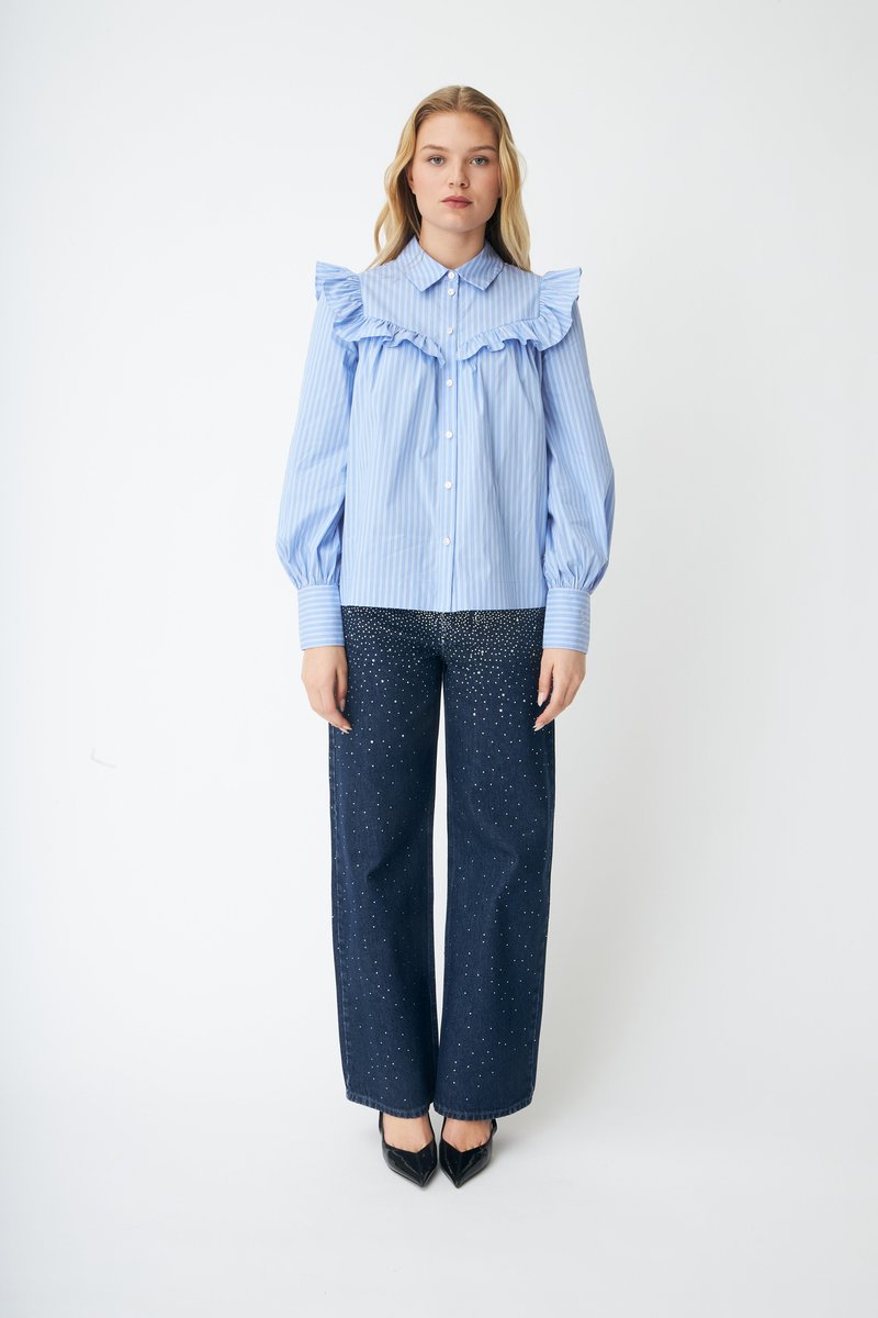 Kvinne iført en blåstripet bluse med volanger og vide, mørke jeans dekorert med små hvite prikker, stående foran en ensfarget hvit bakgrunn.