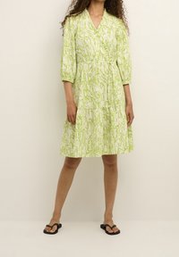 Grünes gemustertes Kleid mit V-Ausschnitt, Dreiviertelärmeln und einem gestuften Rock, aus strukturiertem Stoff. Getragen mit schwarzen Flip-Flops.