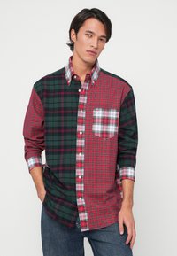 Flanellhemd mit einem halb grünen, halb roten Design und verschiedenen Karo-Mustern. Lange Ärmel, Button-Down-Kragen und eine Brusttasche.