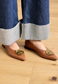 Pointed-toe flat shoes in brown leather with a gold metallic accent. Features a low block heel and is paired with frayed hem denim.