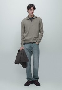 Junger Mann trägt einen taupefarbenen Pullover mit Viertelreißverschluss, helle blaue Jeans und dunkle Schuhe und hält eine dunkle Jacke vor einem schlichten, hellen Hintergrund.