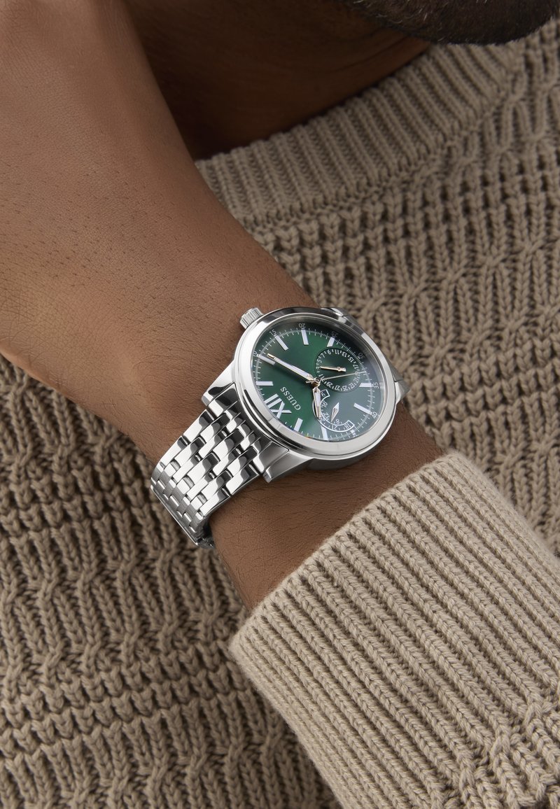 Hõbedane randme kell rohelise kellapaneeli, ümmarguse näo, valgete tunni markerite ja roostevabast terasest punutud käevõruga. Kandja käel, millel on kootud kampsun.