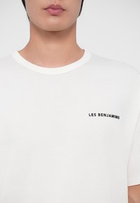 Gros plan d'une personne portant un t-shirt blanc avec le texte "LES BENJAMINS" imprimé en noir sur le côté gauche de la poitrine.