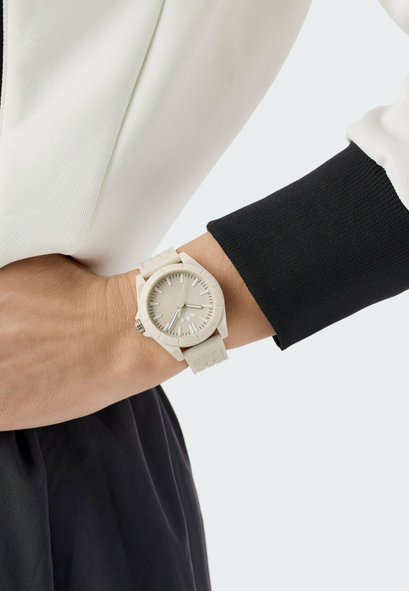 Montre beige clair avec un cadran texturé et des index clairs, fixée par un bracelet en silicone assorti, portée sur un poignet avec une manche noire.