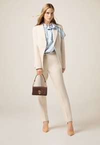 Completo beige su misura con giacca aderente e pantaloni dritti. Blusa a righe blu chiaro e borsa marrone con dettagli dorati. Tacchi nudi.