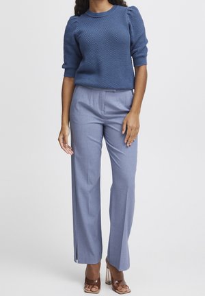 Vrouw draagt een blauw gebreide korte trui met pofmouwen, lichtblauwe rechte broek met hoge taille en bruine open-gespen hakken, staand tegen een witte achtergrond.