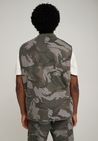 Chaleco con estampado de camuflaje en tela verde y gris, con cuello acanalado y corte recto, combinado con una camisa blanca de manga corta.