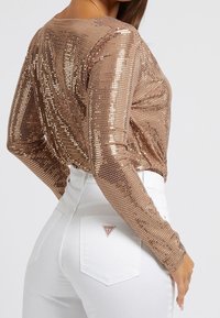 Rose gold sequin long-sleeve top, fitted design, light shimmer, paired with high-waisted white denim jeans featuring logo patch on back pocket.