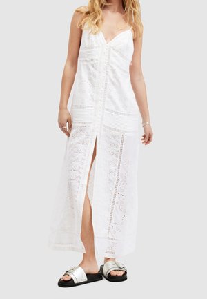 Nainen pukeutuneena valkoiseen pitsiseen maksimekkoon, jossa on etuhalkaisija, yhdistettynä hopeisiin solkisandaaleihin vaaleanharmaalla taustalla.