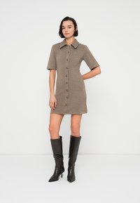 Καφέ πλεκτό φόρεμα με γιακά στο λαιμό, κοντά μανίκια και κουμπιά στο μπροστινό μέρος· συνδυασμένο με μαύρες δερμάτινες μπότες μέχρι το γόνατο.