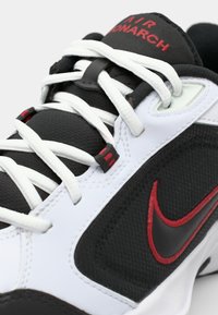 Sort og hvid atletiksko med røde accenter. Har mesh- og syntetiske materialer, tykke snørebånd og et fremtrædende Nike-logo på siden.