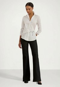 Ženska v beli srajci z gumbi in stransko verižico, črnih hlačah s širokimi nogavicami in črnih visokih petah, stoji.