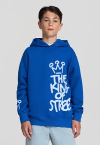 Niebieska bluza z kapturem wykonana z miękkiego materiału, z dużym białym nadrukiem z napisem "KRÓL ULICE" oraz wzorem korony. Z przodu kieszeń typu kangur.