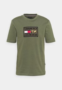 Camiseta de algodón color verde oliva con un parche frontal con un logo, acentos en rojo y blanco, y bordes negros. Cuello redondo y mangas cortas.