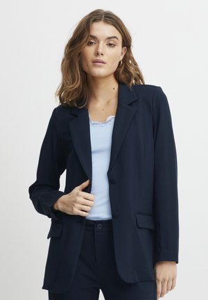 Blazer marine à revers, avec une fermeture à un bouton et deux poches avant. Porté par-dessus un haut bleu clair, présentant un motif texturé en côtes.