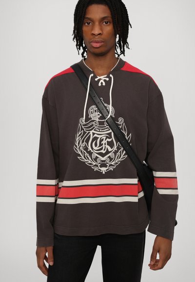 Camisa marrón de manga larga con acentos rojos en los hombros y rayas horizontales. Presenta un diseño gráfico de escudo y un escote con cordones. Banda negra en el hombro.